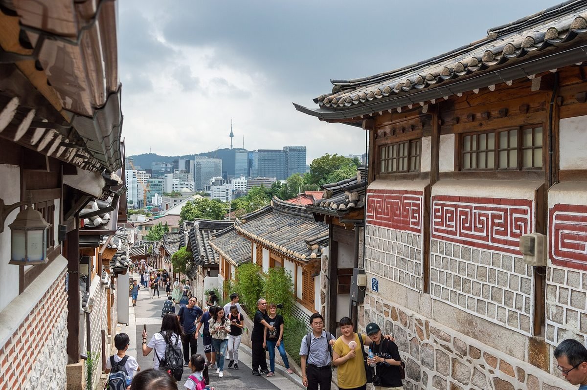 한옥 처마 사이로 남산서울타워와 도심 빌딩이 보이는 북촌로11길 내리막에 관광객들이 줄지어 걷는 모습