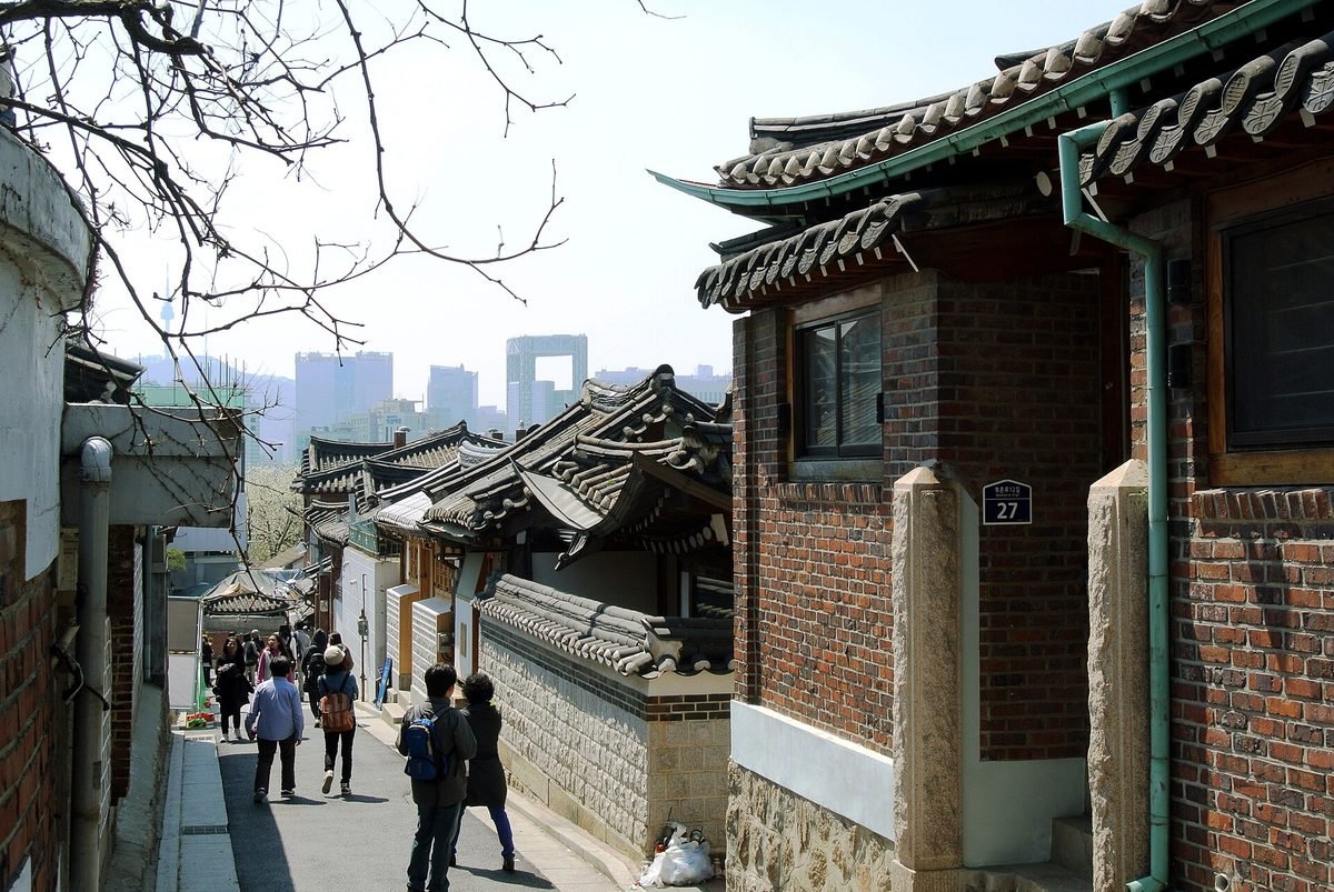 붉은 벽돌 한옥과 기와 담장 사이 좁은 골목을 배낭 멘 관광객들이 걸어 내려가고 멀리 고층 빌딩이 보이는 북촌로12길