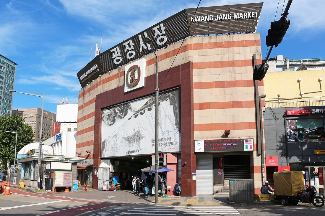 '광장시장 KWANG JANG MARKET' 간판이 걸린 붉은 줄무늬 시장 출입구 건물과 앞쪽 횡단보도