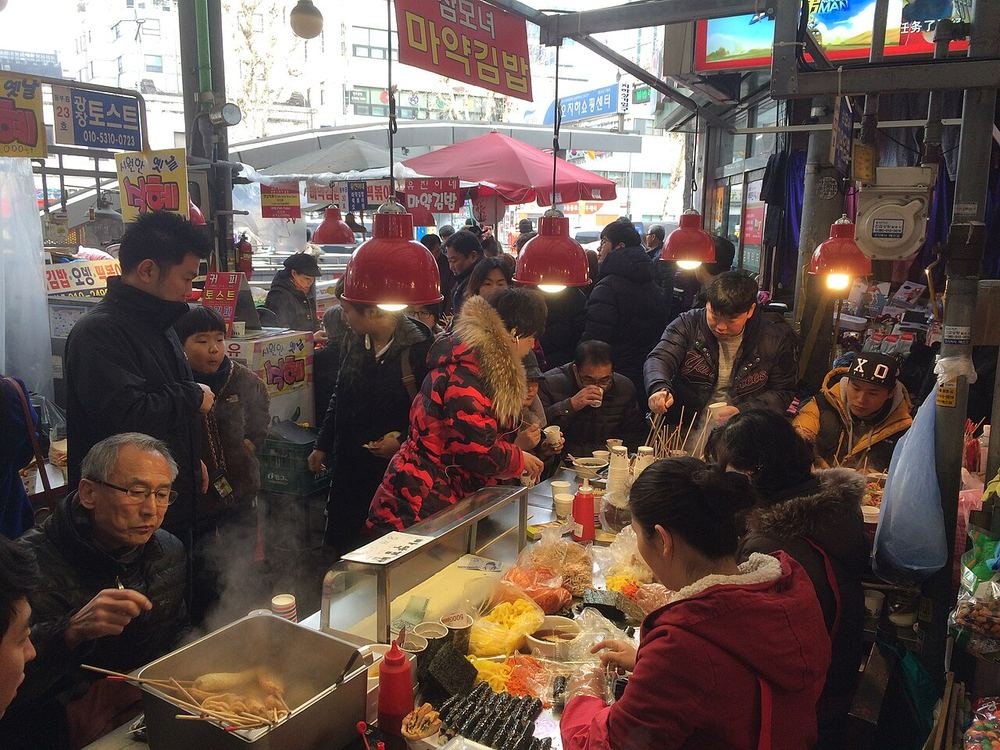 빨간 조명 아래 노점 앞에 둘러앉은 손님들 앞에서 상인이 꼬마김밥을 말고 있고, 위에 마약김밥 간판이 걸린 모습