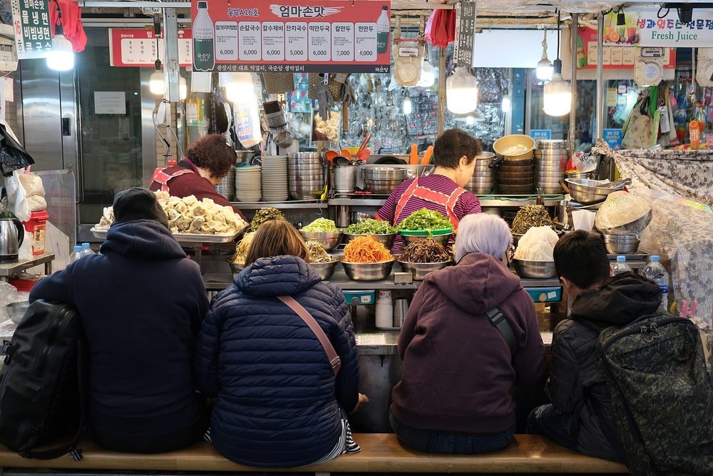 나물 그릇과 만두가 놓인 노점 앞 긴 나무 의자에 나란히 앉아 음식을 먹는 손님들과 그 너머의 상인들