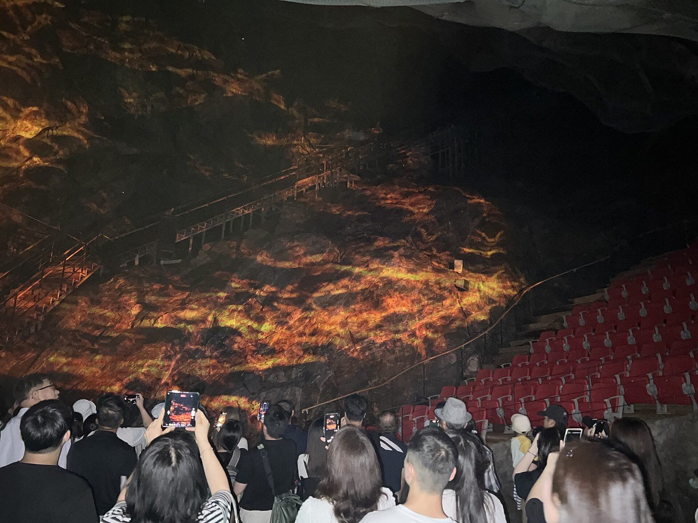 붉은 객석이 놓인 동굴 공간에서 관람객들이 휴대전화로 바위벽에 비친 주황빛 영상을 찍는 모습