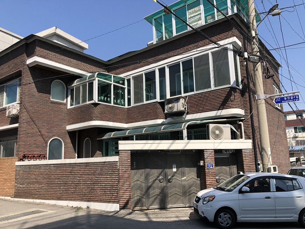 2층 붉은 벽돌 주택의 2층에 유리 베란다가 달려 있고 대문 앞에 흰색 소형차가 서 있는 모습
