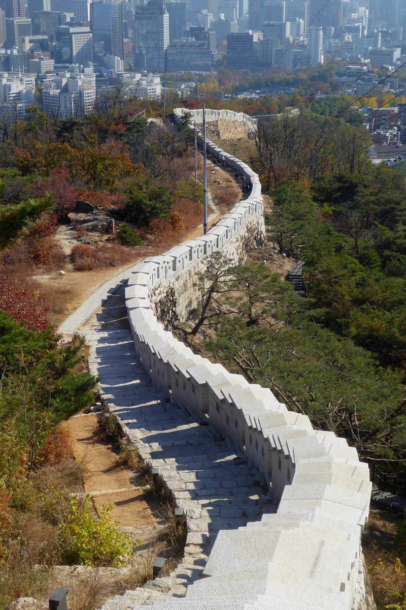 인왕산 능선을 따라 굽이치며 내려가는 흰 성곽과 옆의 돌계단 길, 멀리 보이는 도심 빌딩