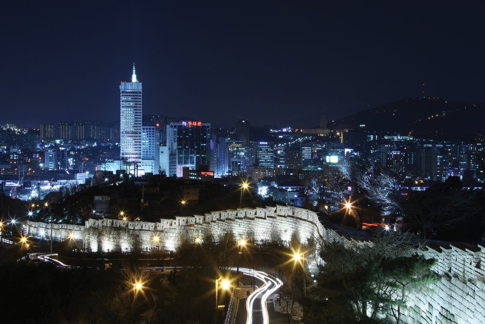 밤에 조명을 받은 낙산 성곽과 그 뒤로 불이 켜진 고층 빌딩, 아래쪽 도로의 차량 불빛 궤적